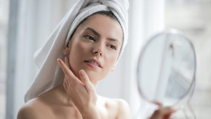 selective focus portrait photo of woman with a towel on head looking in the mirror - Skin Care Routines
