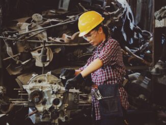 woman wears yellow hard hat holding vehicle part - huge gender pay gap in trades industries today