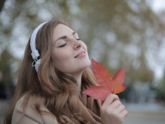 young satisfied woman in headphones with fresh red leaf listening to music with pleasure while lounging in autumn park