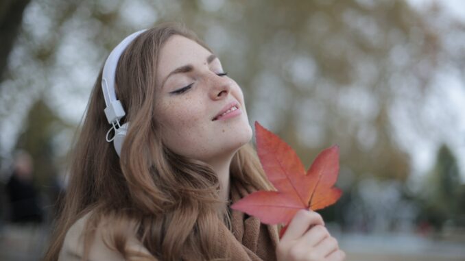 young satisfied woman in headphones with fresh red leaf listening to music with pleasure while lounging in autumn park